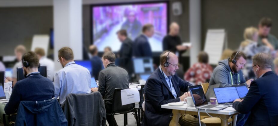 Ihmiset istuvat pöytien ääressä. Pöydillä on kannettavia tietokoneita ja monella ihmisellä on kuulokkeet päässään. Taustalla seisoo lisää ihmisiä.